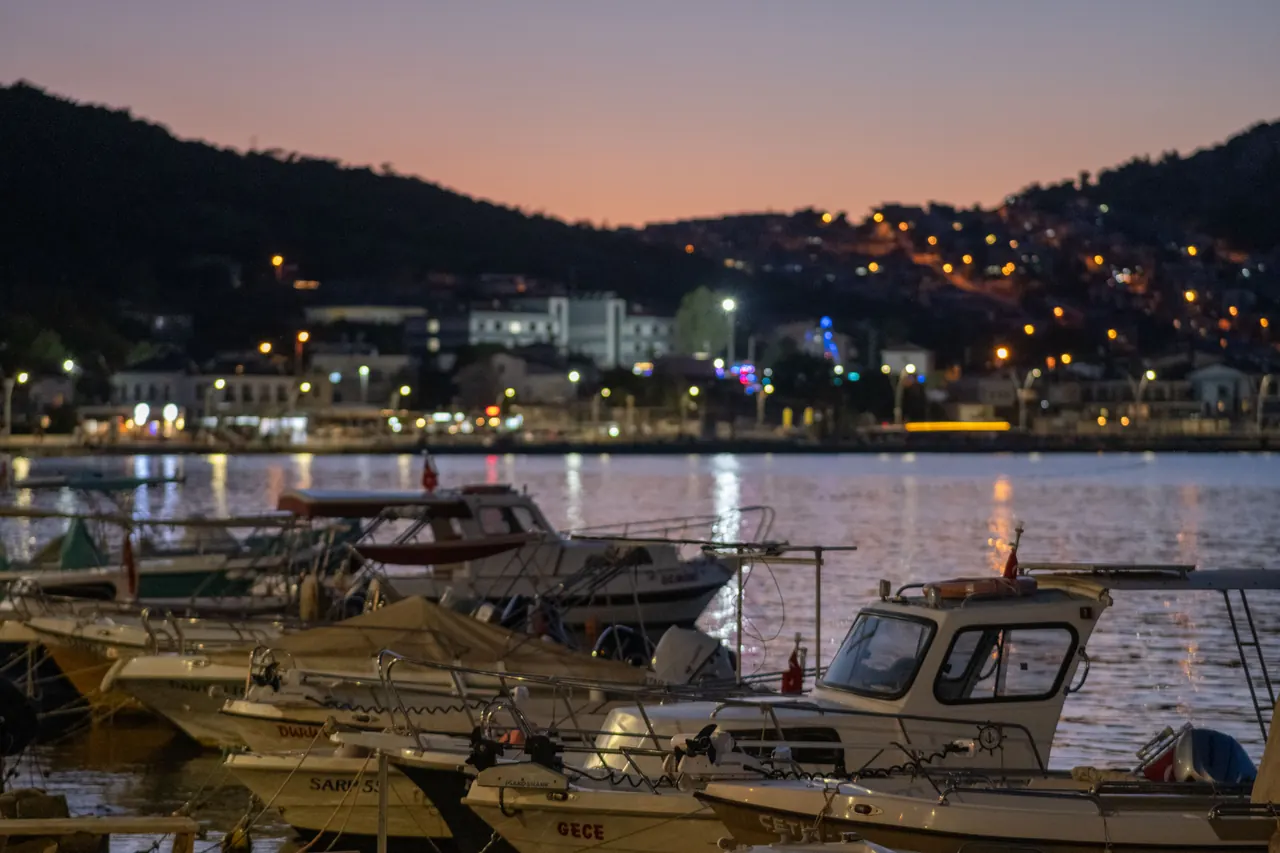 Boats resting at Foça harbor during a vivid sunset with city lights sparkling in the distance.