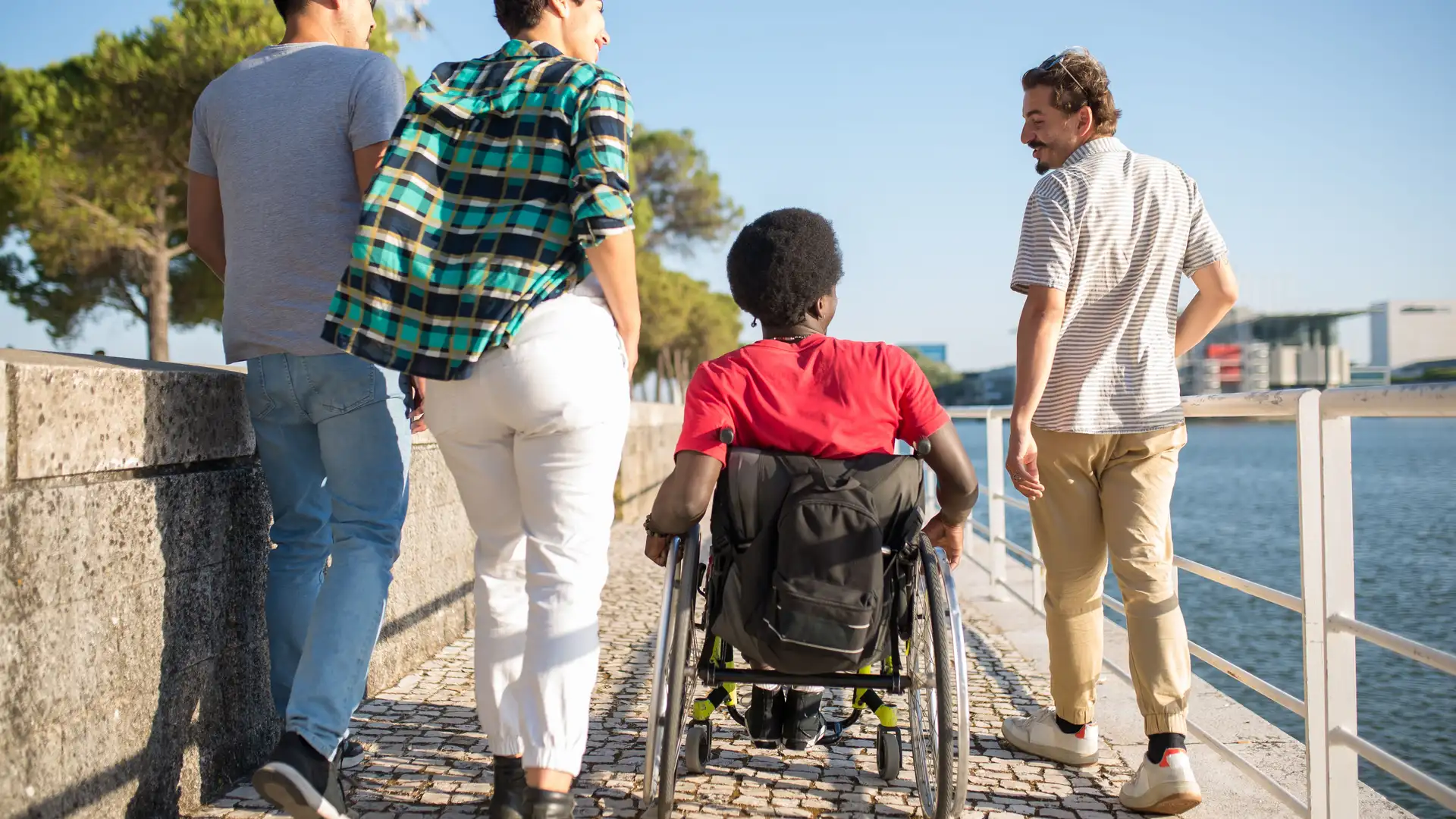 A group of friends walking together along a riverside path, one of them using a wheelchair