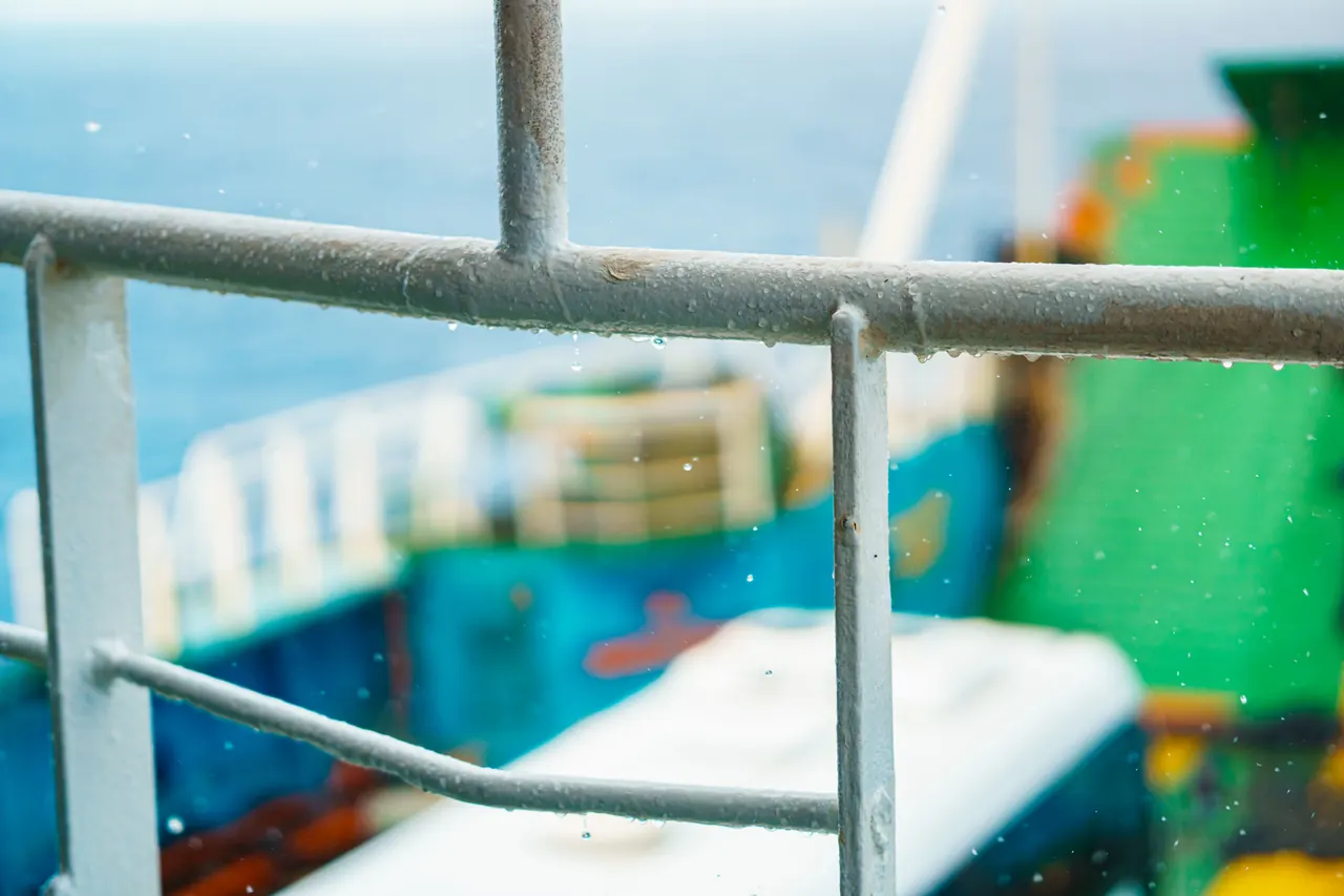 Close-up of a metal railing with a blurred cargo ship in the background over the ocean.