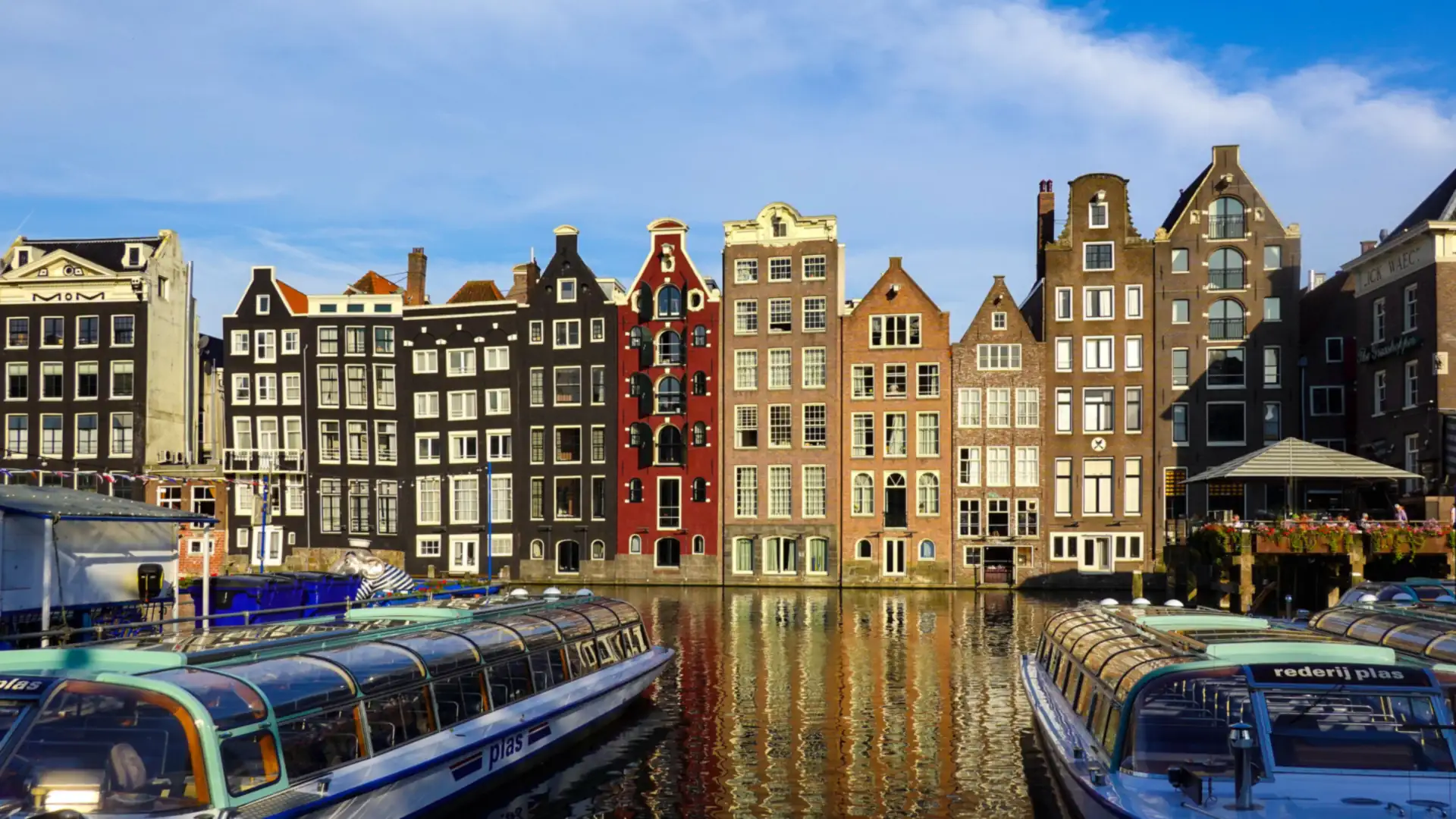 Tall, narrow historic canal houses in Amsterdam reflected in the water, with glass-roofed tour boats moored in the foreground