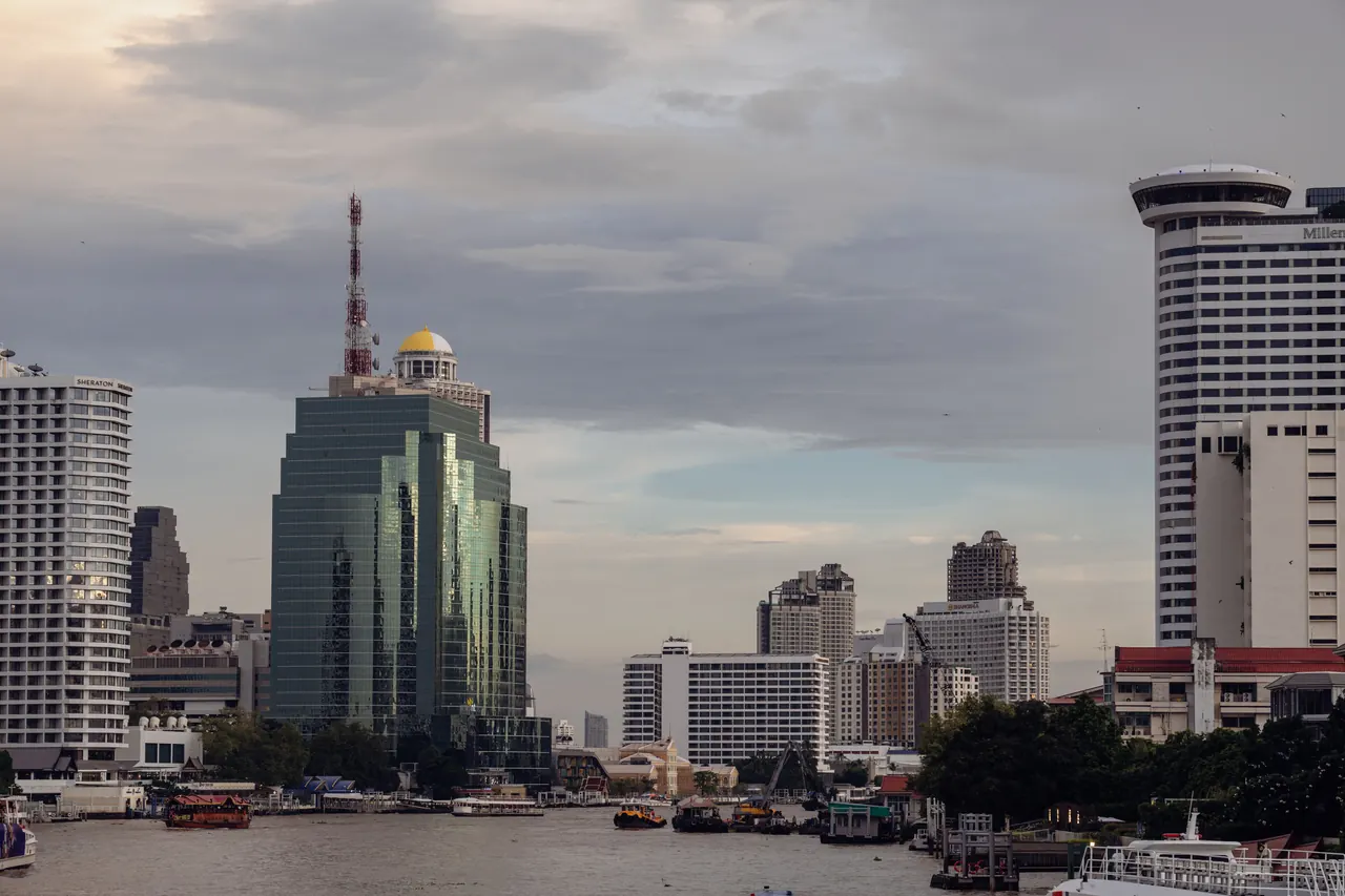 View of Bangkok skyline featuring modern buildings and the Chao Phraya River under a cloudy sky.