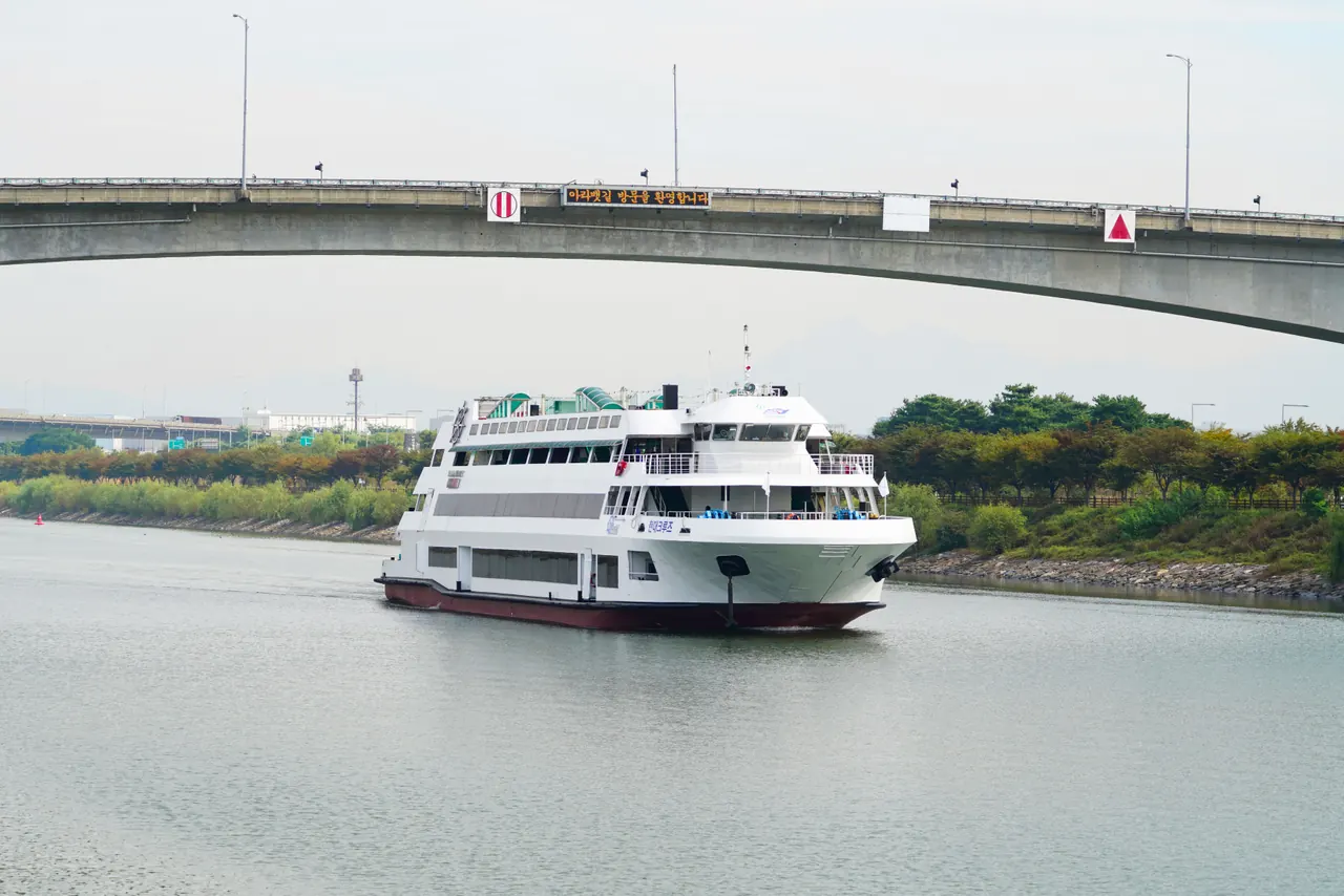 A cruise ship navigates a serene waterway beneath a concrete bridge, set against a lush green landscape.