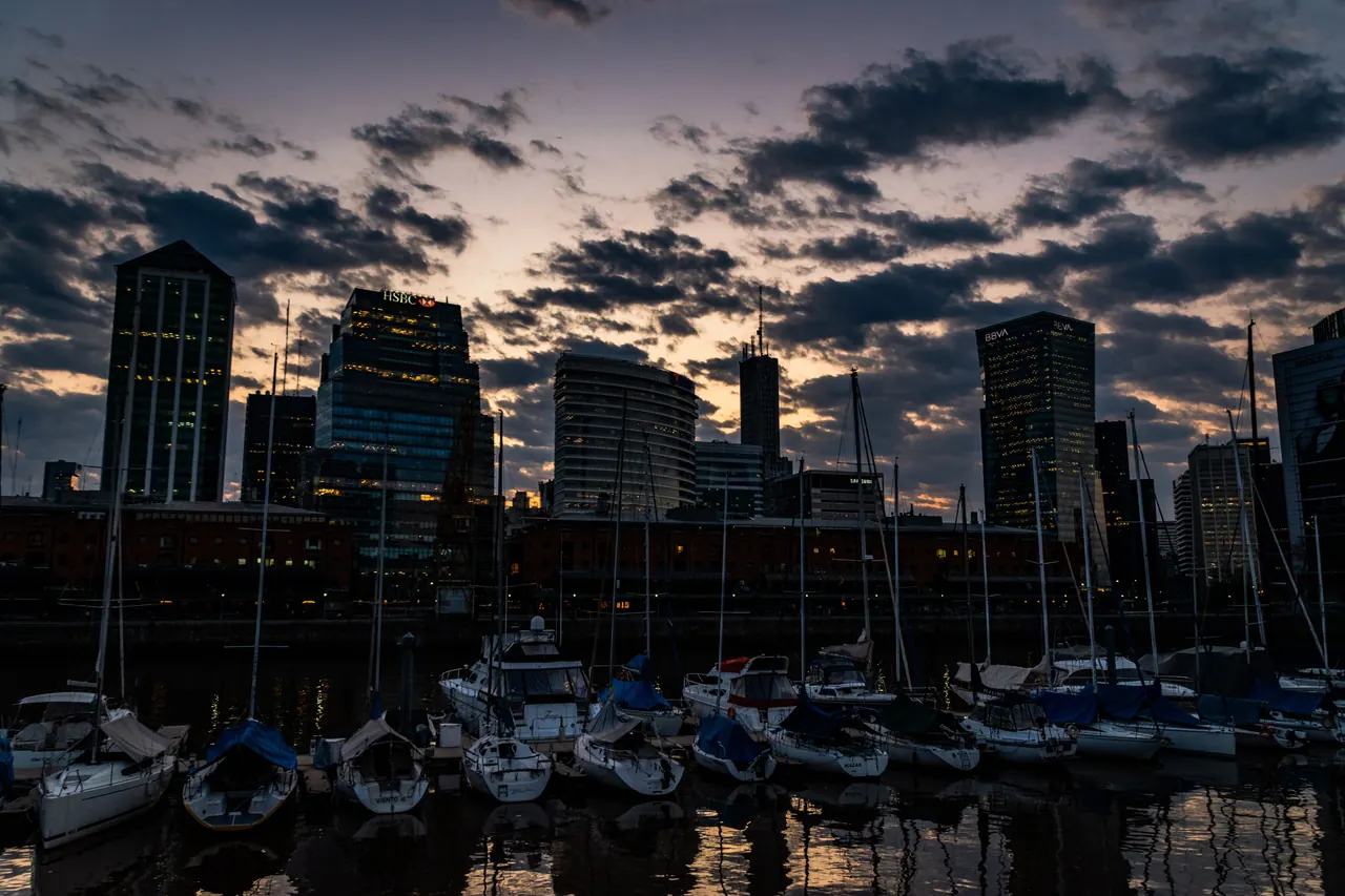 Silhouette of city skyline at dusk with boats moored in the marina reflecting on calm water.