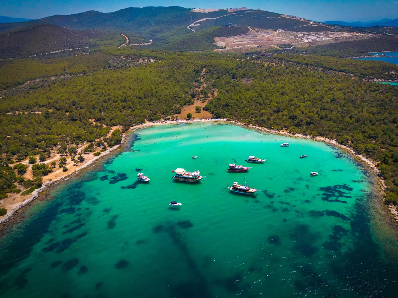 A stunning aerial view of a turquoise bay lined with lush forest, featuring several yachts anchored along the shore.