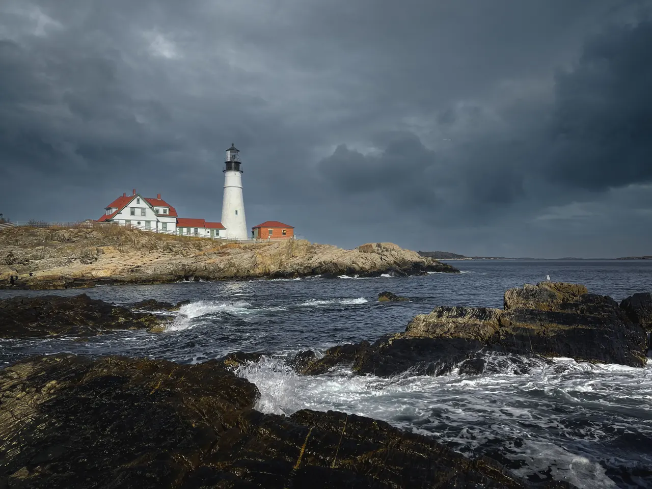Lighthouse and houses located on shore with rocks washed by foamy waves in gloomy overcast day