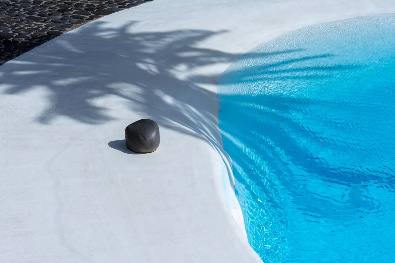 Minimalist scene of a poolside with a tranquil black stone and palm tree shadow on concrete.