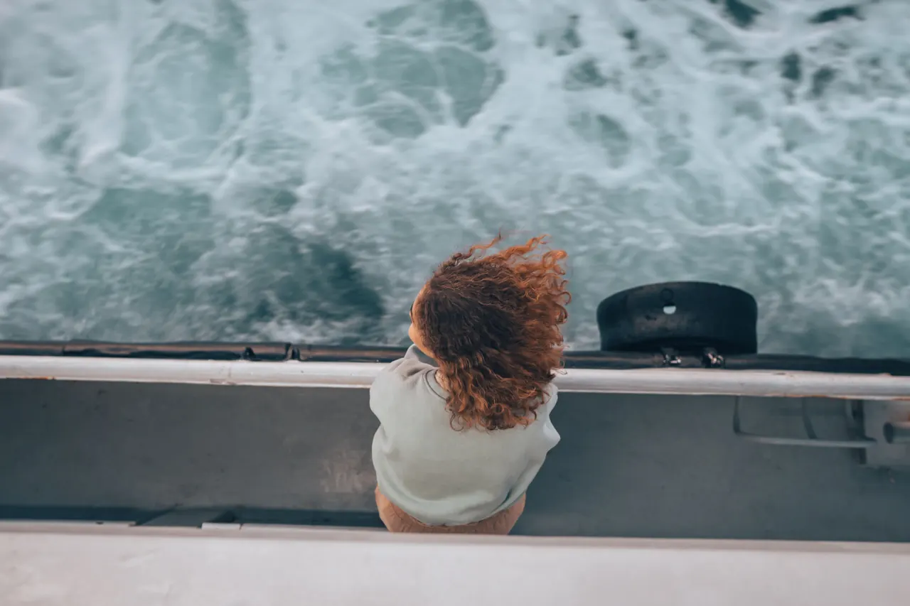 A woman with curly hair leans over a boat, enjoying the sea breeze and ocean waves.
