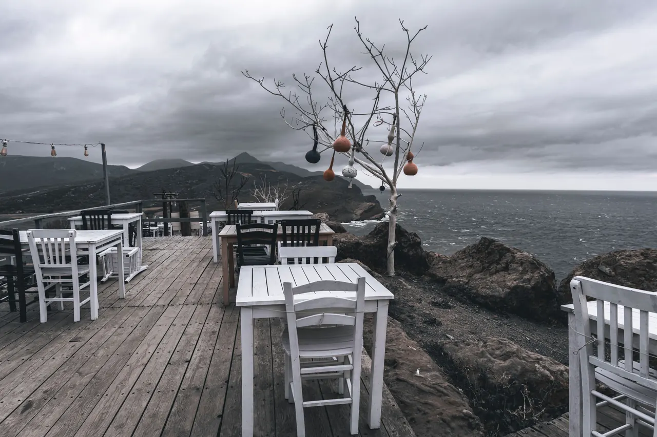 Outdoor terrace overlooking a sea coast with white wooden tables under an overcast sky.