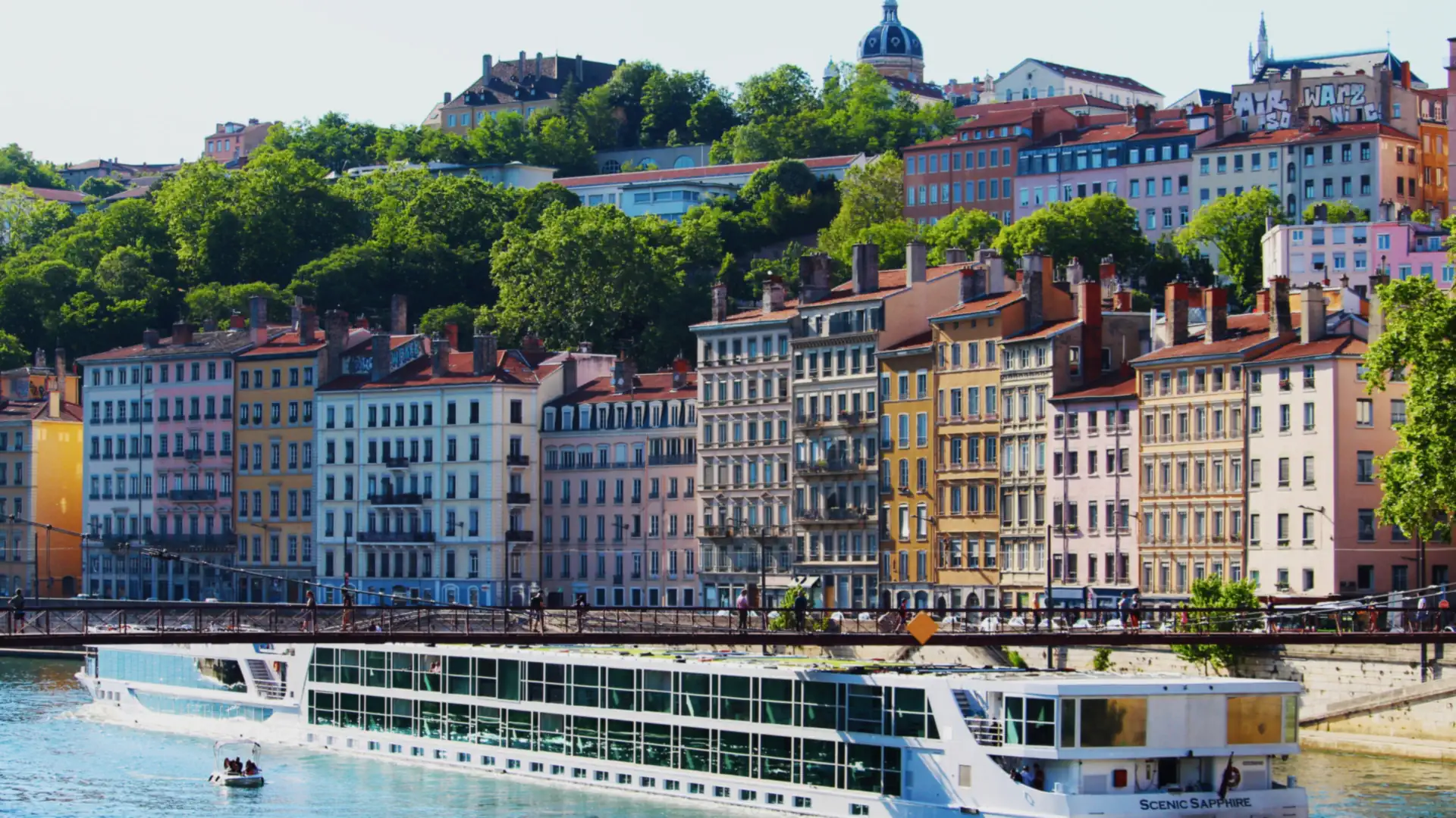 A long river cruise ship moored along a riverbank lined with colorful historic buildings and a hilltop basilica in Lyon, France