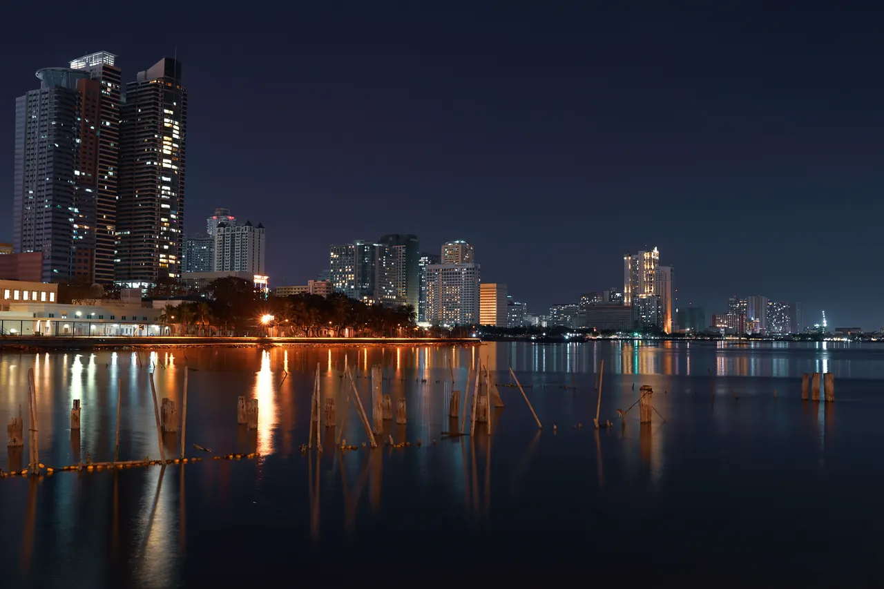 Illuminated skyscrapers reflected on the waterfront in Manila cityscape at night.