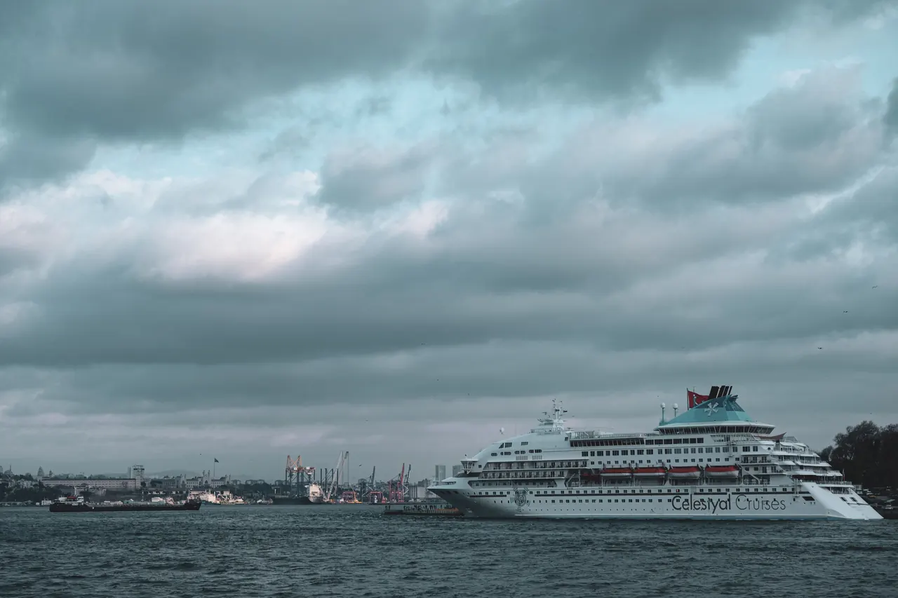 A large cruise ship anchored at a busy port with a dramatic cloudy sky overhead, creating a moody maritime scene.