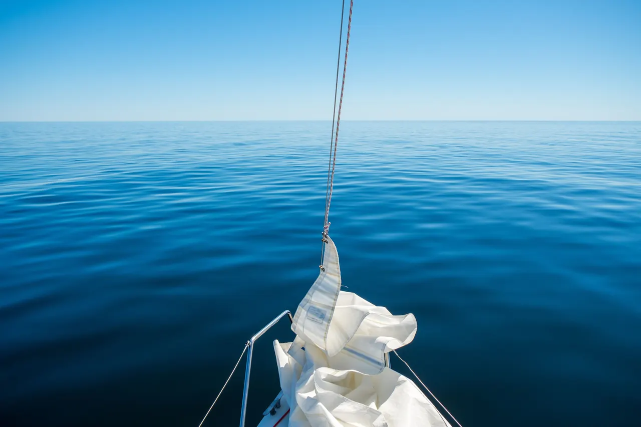 Tranquil scene of a sailboat on a calm, blue ocean under a clear sky, ideal for travel and relaxation themes.
