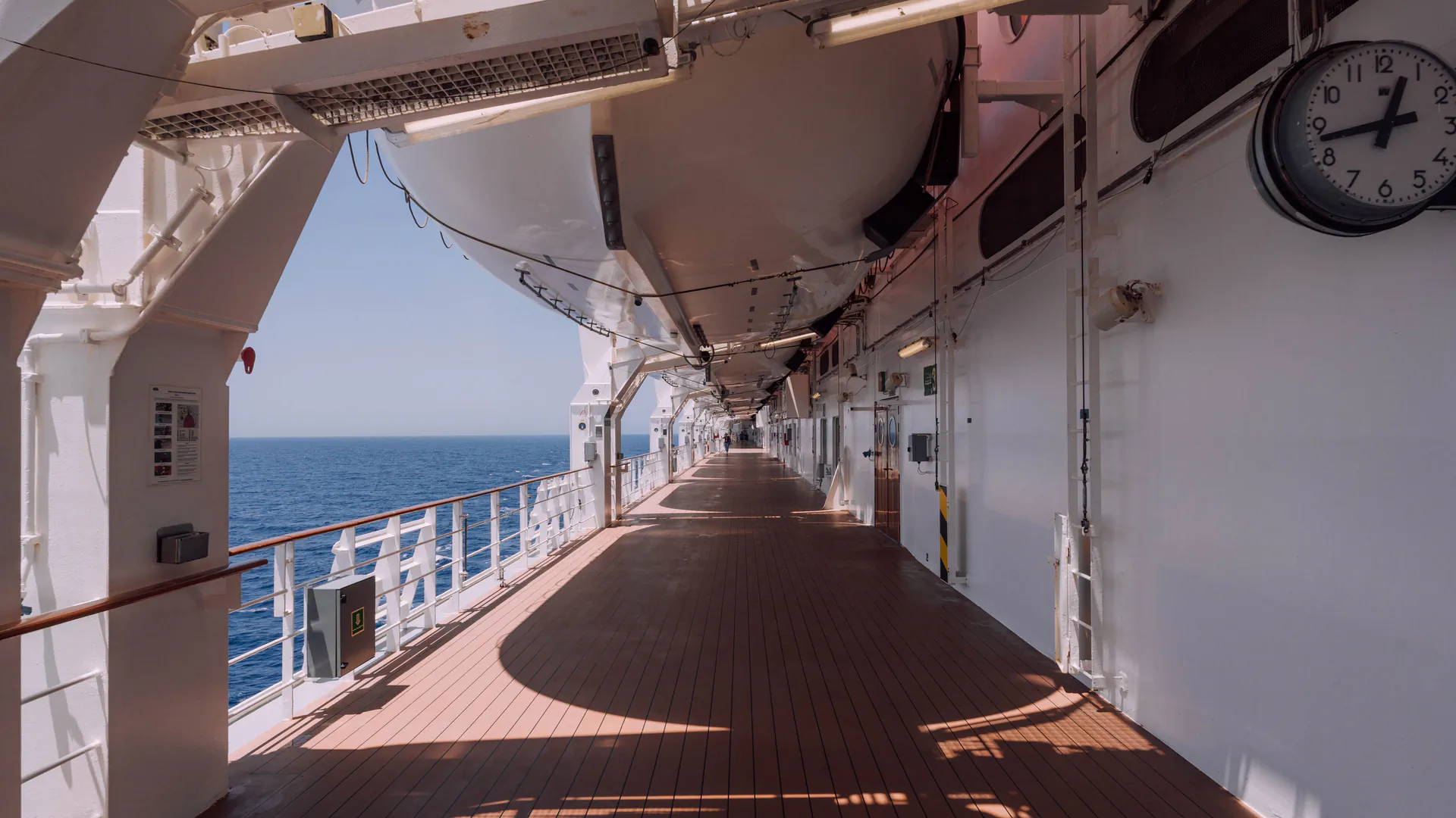 An empty teak promenade deck on a cruise ship, looking along the rail toward a calm sea