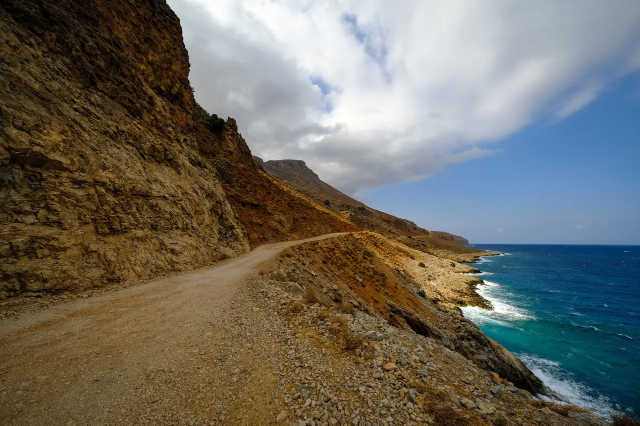 Picturesque dirt road along rocky cliffs and blue sea, under a partly cloudy sky.
