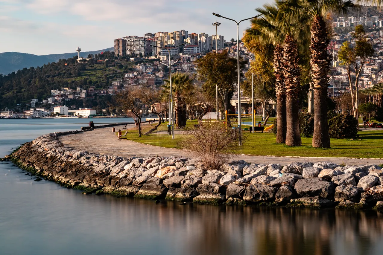 Serene coastal landscape of İzmir with city buildings and lush greenery under a bright sky.