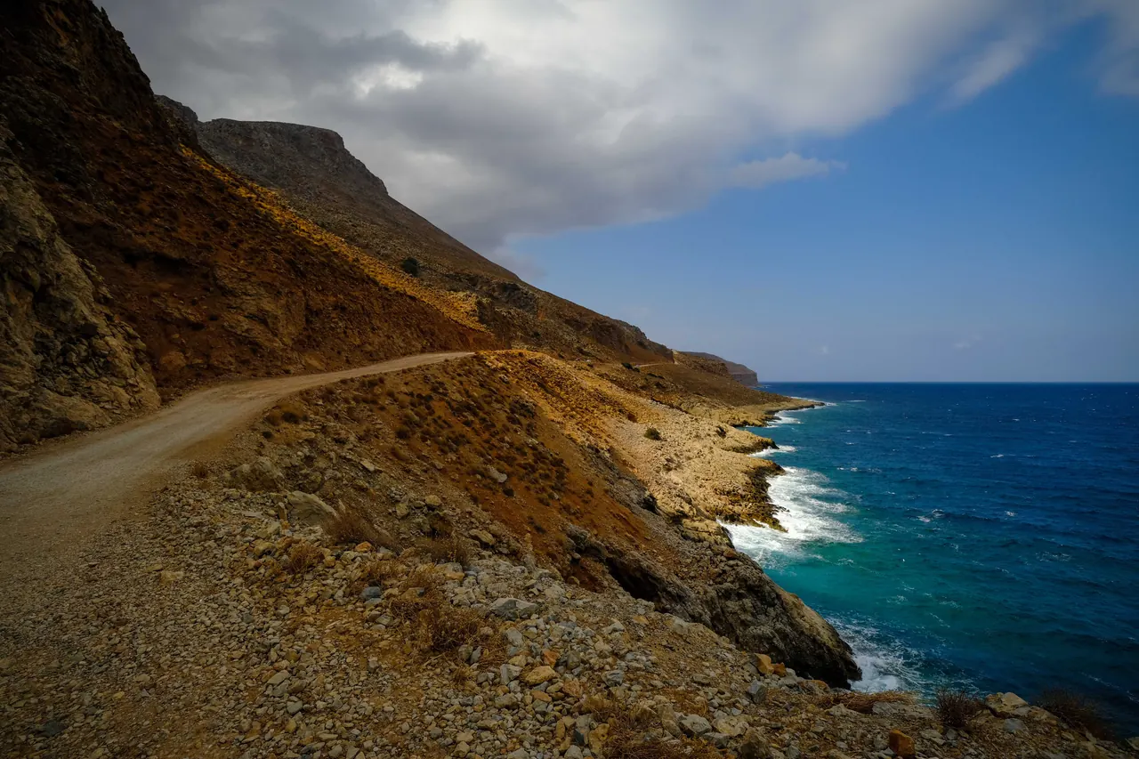 Rustic coastal road winding along a rocky cliff with ocean views under a blue sky.