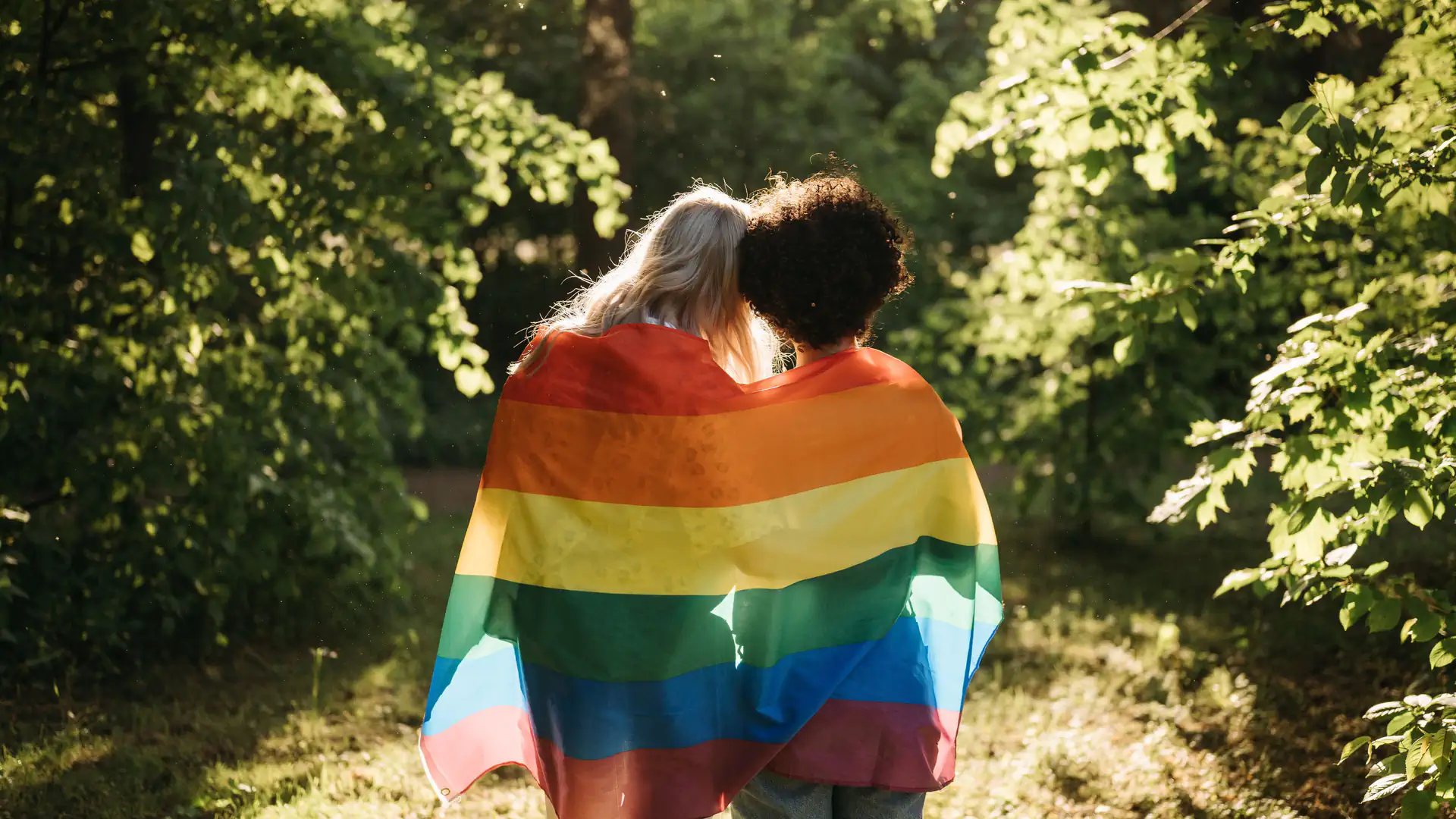 Two people wrapped together in a rainbow flag walking through a sunlit park