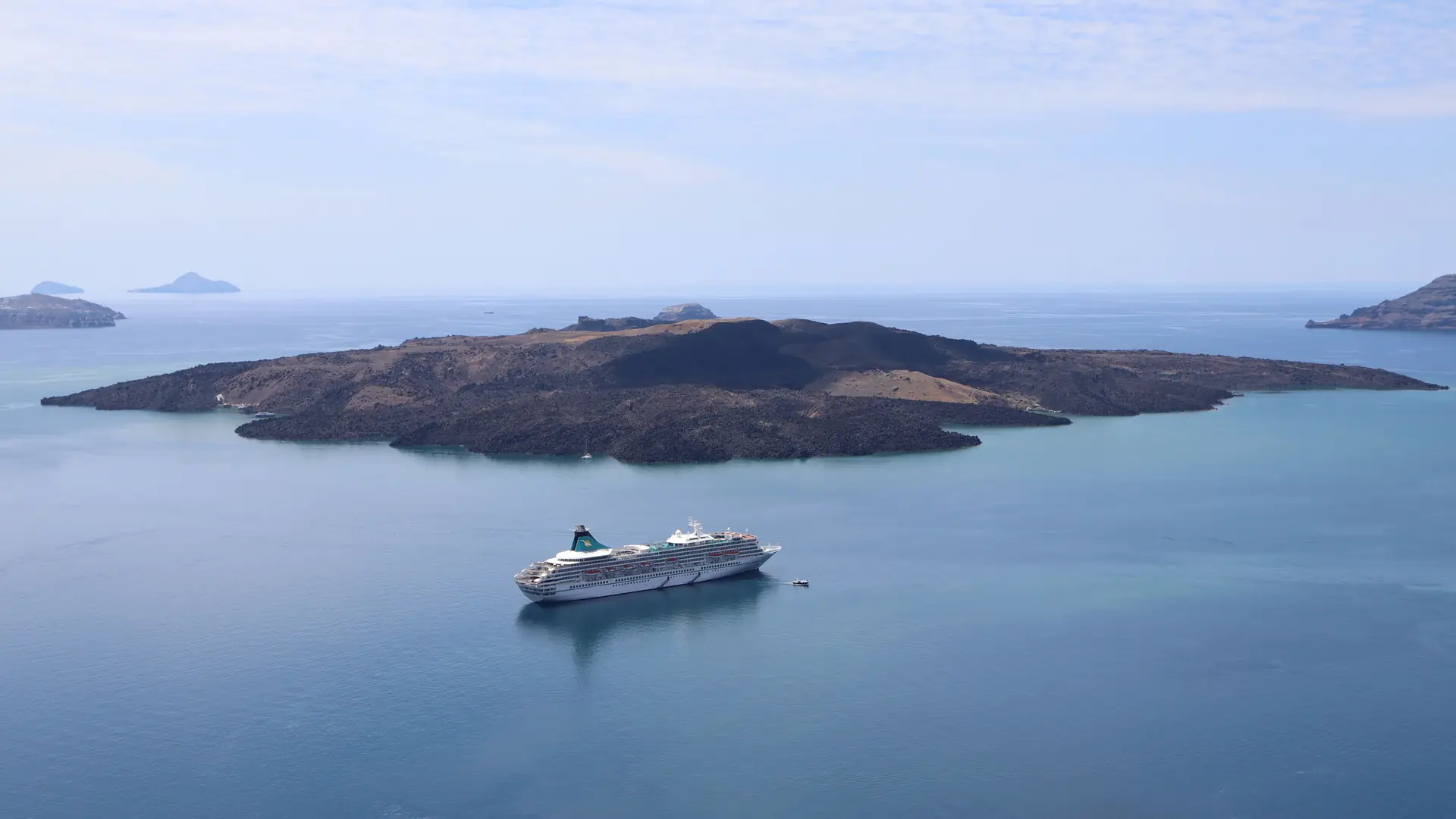 A large cruise ship sailing on calm blue water past a dark volcanic island in the Aegean