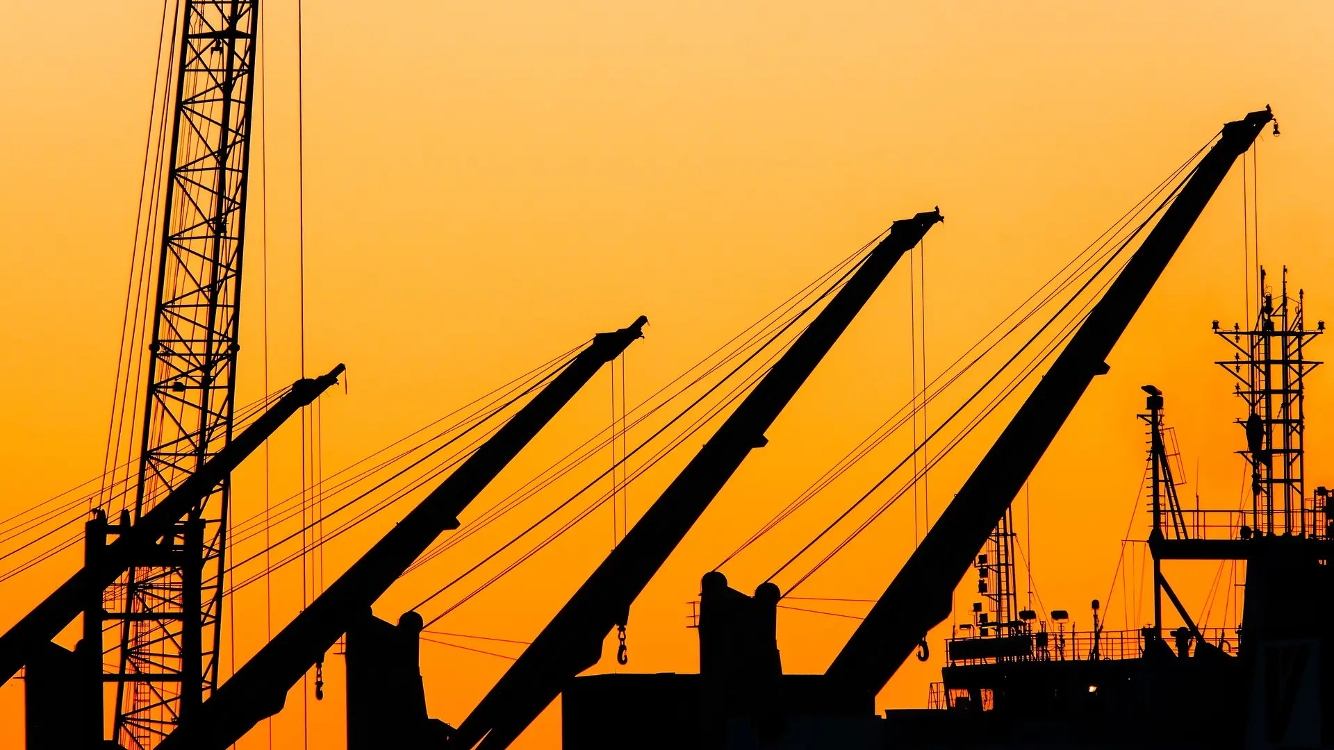 Silhouetted shipyard cranes against a vivid orange sunset sky