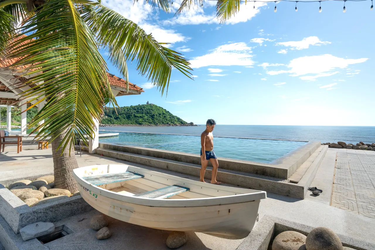Young boy walking near an infinity pool by the ocean in a tropical setting, under a clear blue sky.