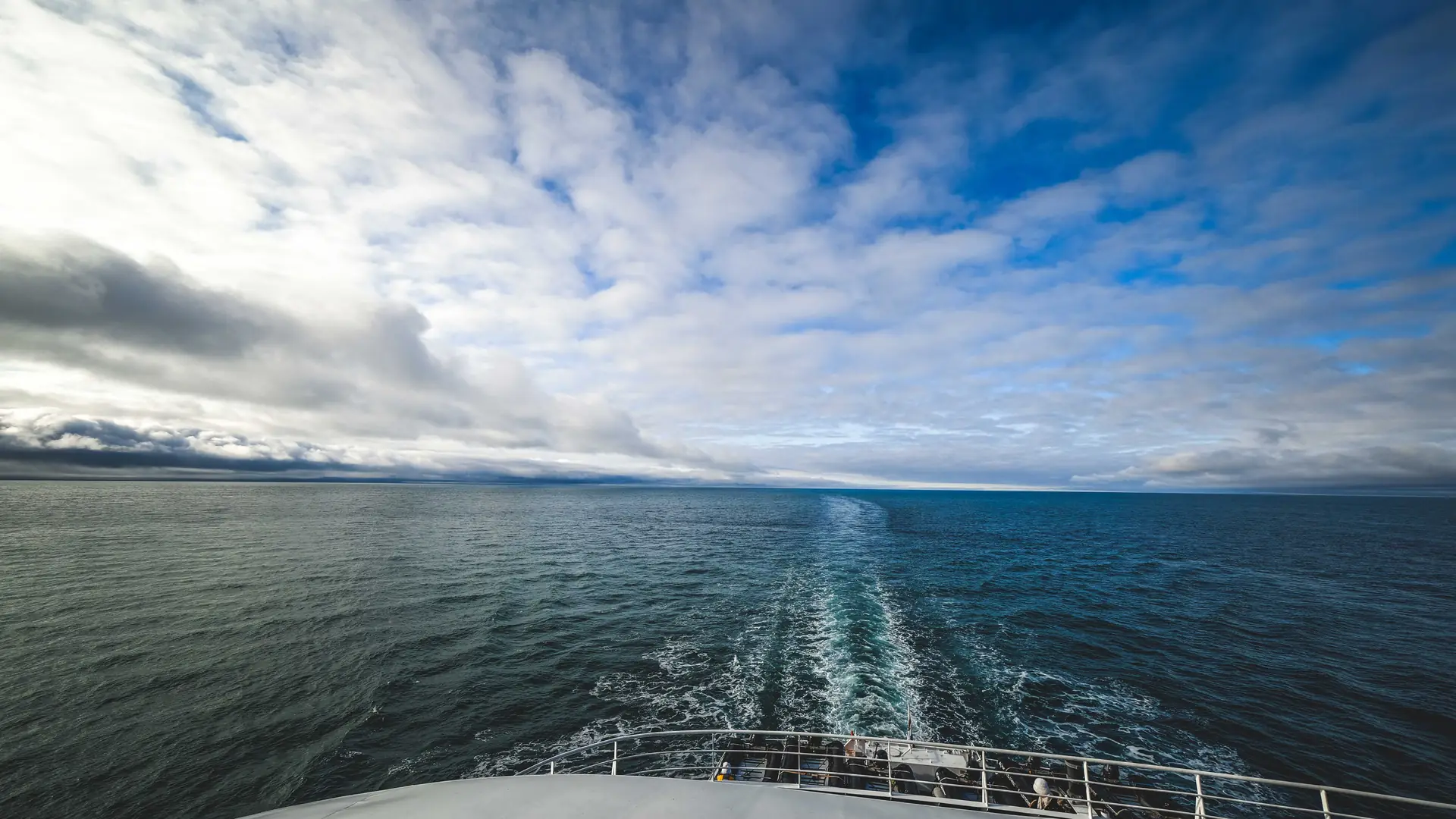 The white bow rail of a ship pointing out over a wide, dark blue ocean under a dramatic sky streaked with clouds