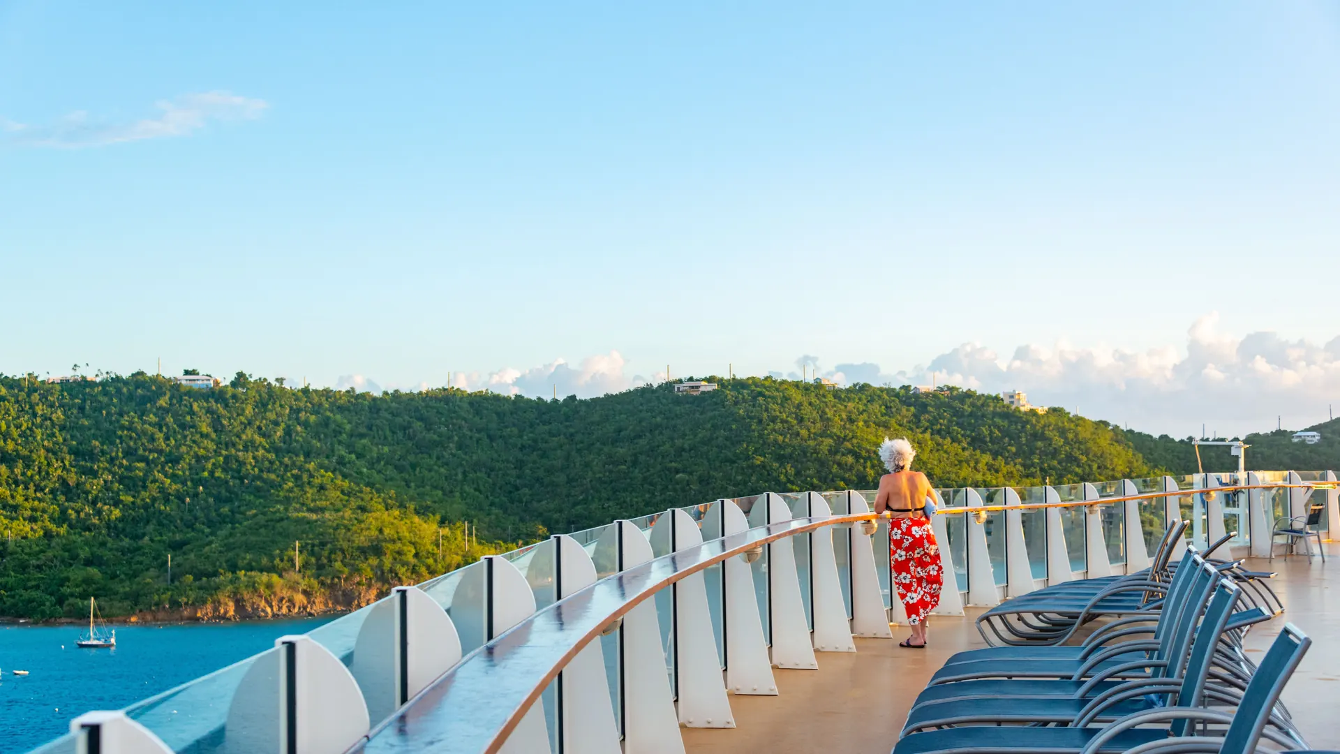 A woman in a red wrap standing alone at the glass rail of a cruise ship deck, looking out at green hills and blue water