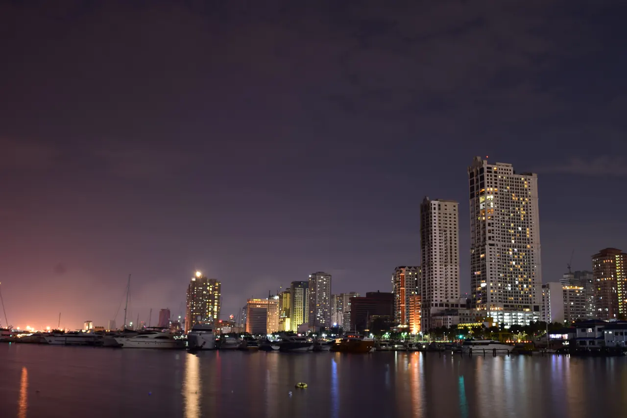 Picturesque scenery of contemporary skyscrapers located on shore of sea with moored boats against dark sundown sky in Manila