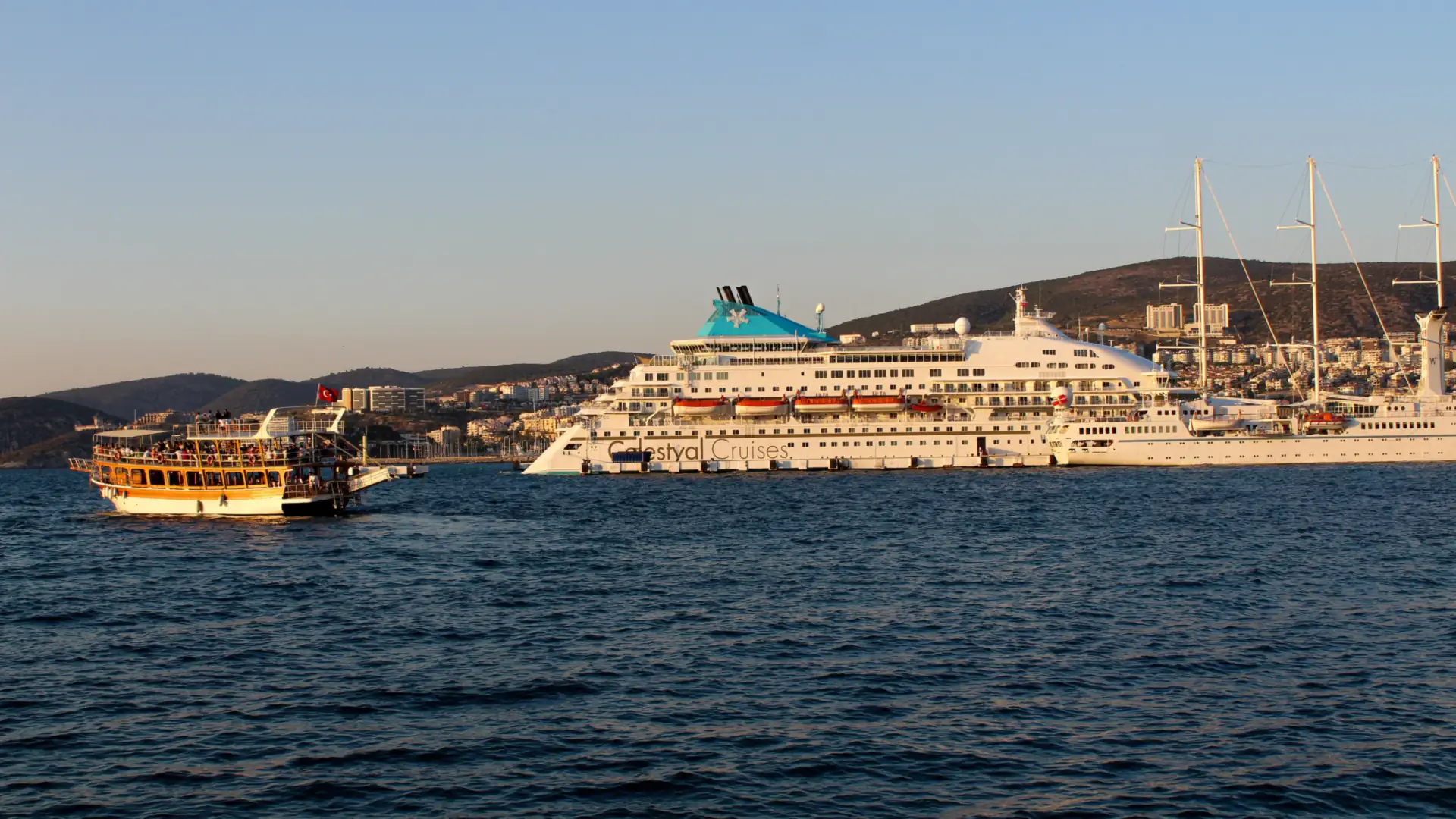 A small tour boat crossing calm blue water in front of a large white cruise ship anchored off a hillside town