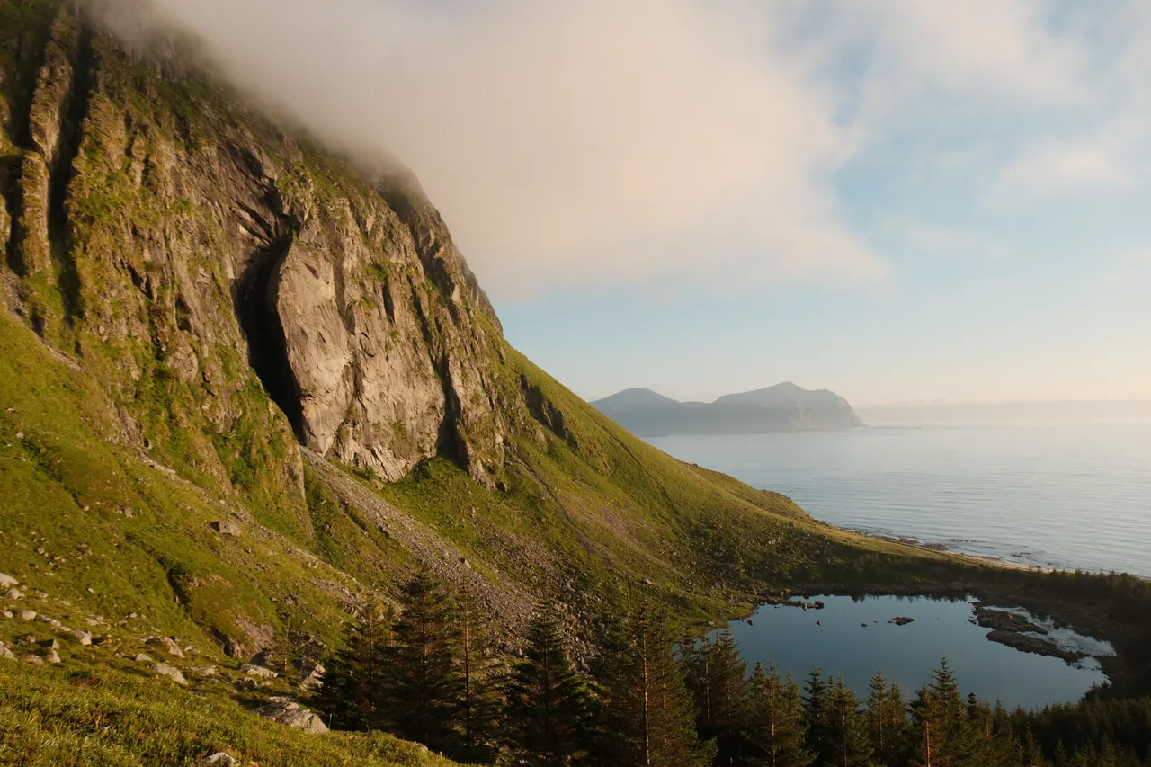 A tranquil alpine scene with towering cliffs, a serene lake, and the ocean beyond under a bright sky.