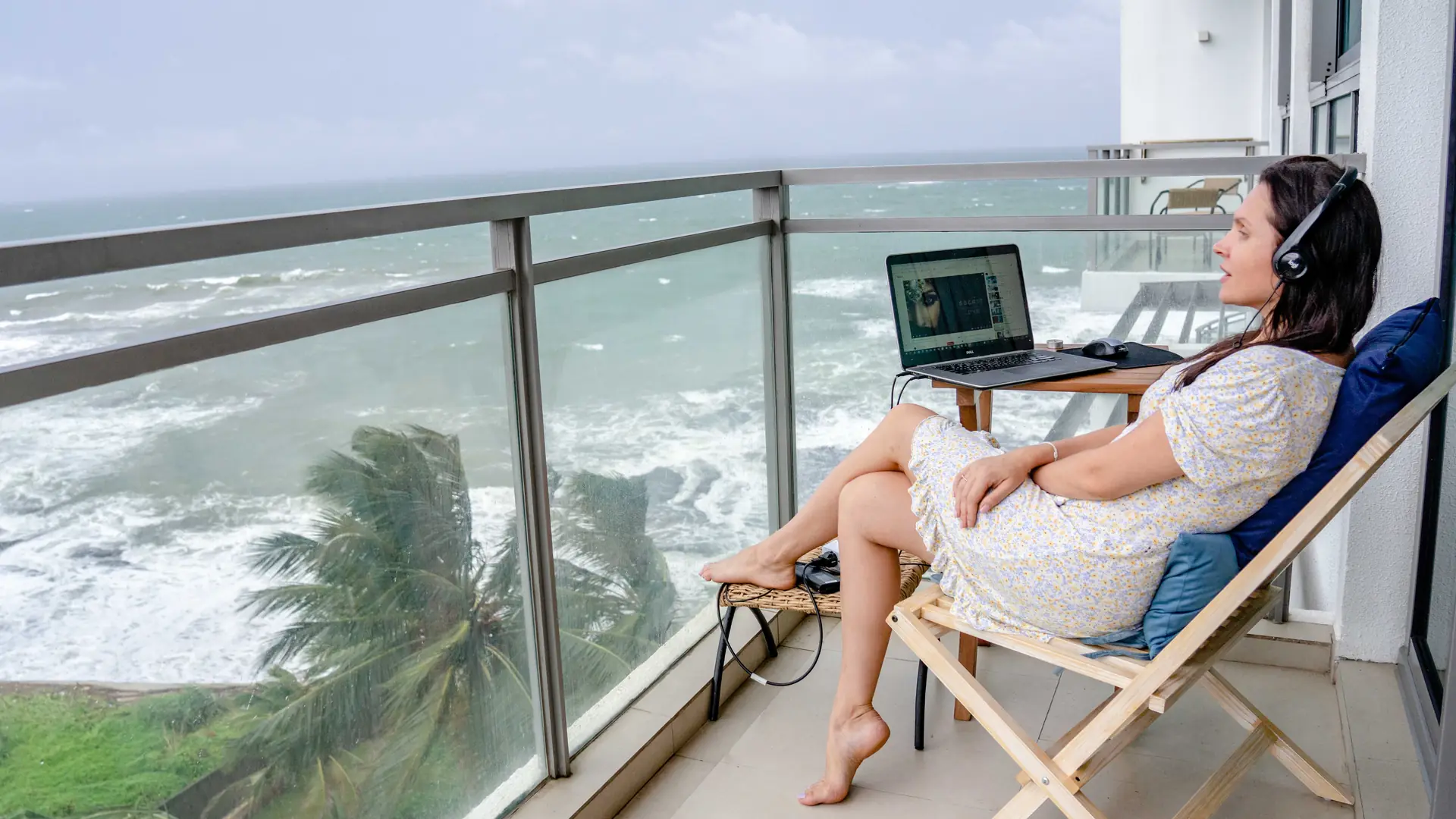 A woman wearing headphones sits in a deck chair on a seaside balcony working on a laptop, with waves and palm trees below